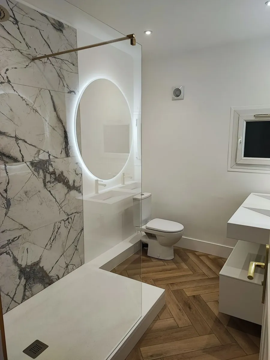 Rénovation de salle de bain complète : douche à l'italienne avec paroi en verre, mur en marbre, miroir lumineux rond, meuble vasque et sol en chevrons